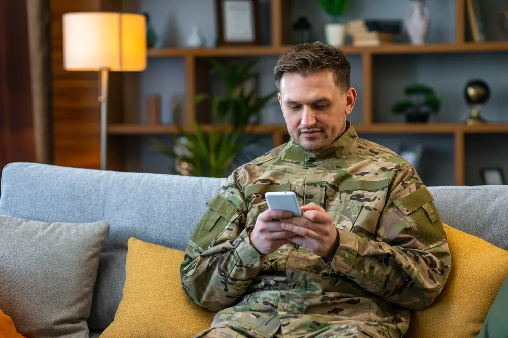 Soldat in Uniform blickt auf sein Smartphone, während er in der Kaserne auf einem Sofa sitzt.