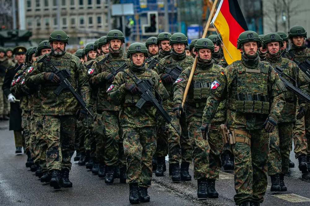 Soldaten der Bundeswehr in Flecktarn-Uniform marschieren mit Gewehren und Deutschlandfahne diszipliniert in Formation.